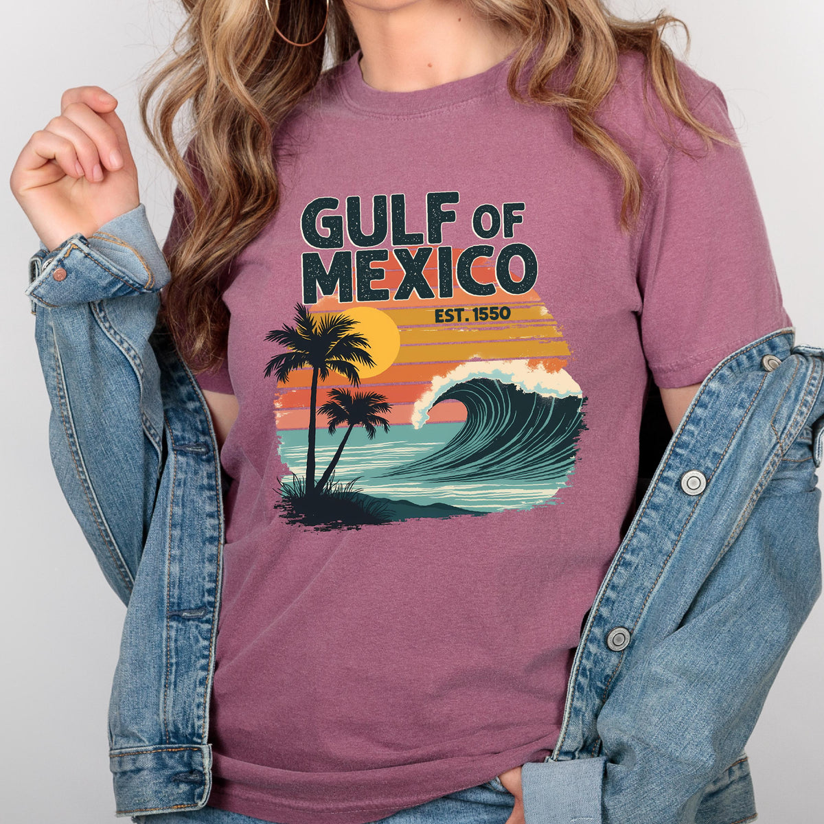 May include: A maroon t-shirt with a retro-style graphic of a wave crashing on a beach with palm trees and a sunset. The text "GULF OF MEXICO EST. 1550" is printed on the shirt.