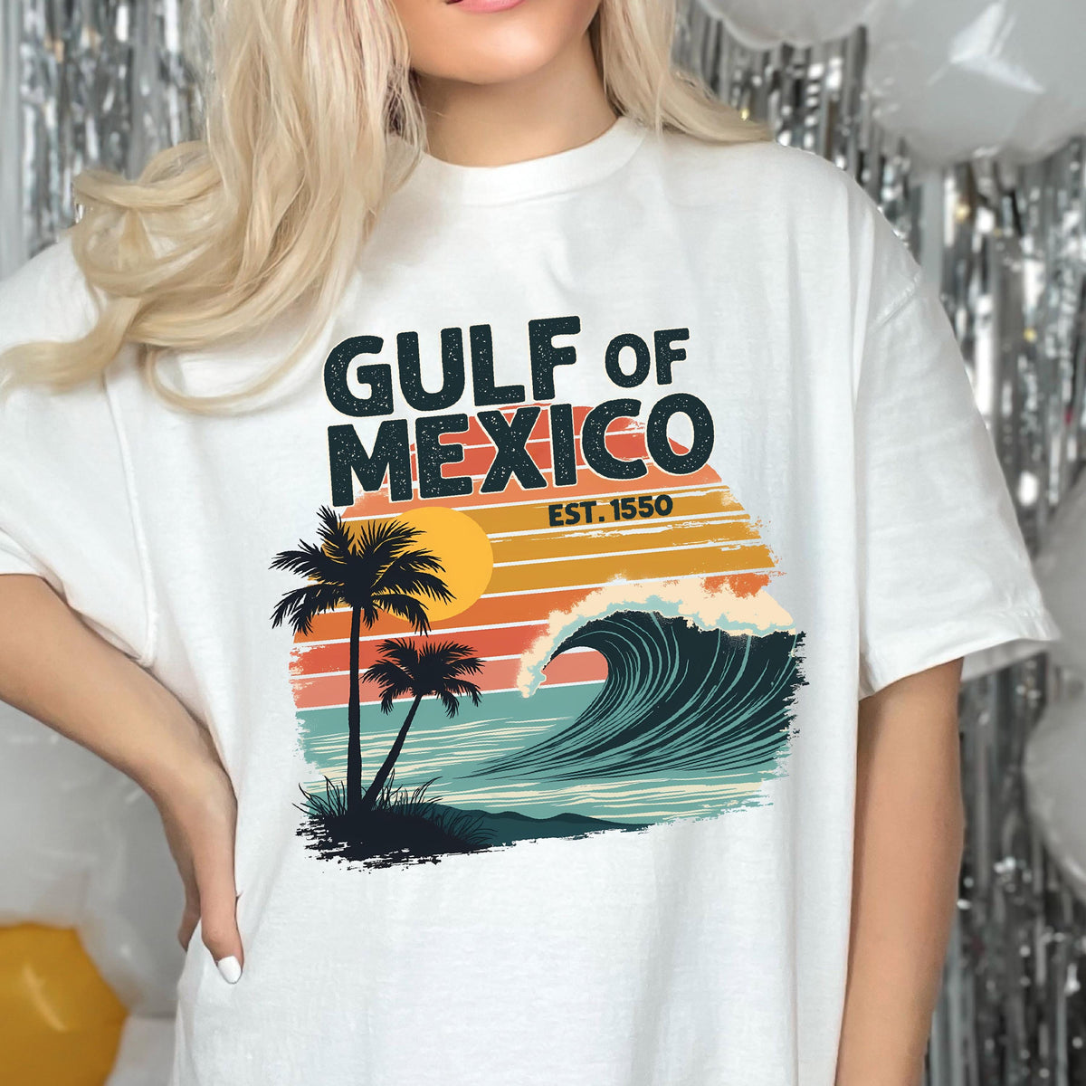 May include: White t-shirt with a graphic design of a wave crashing on a beach with palm trees and the text "Gulf of Mexico Est. 1550".