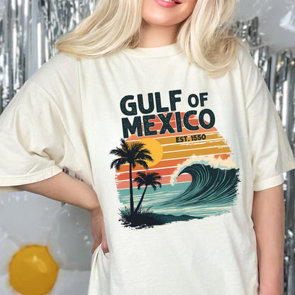 May include: A white t-shirt with a vintage-style graphic design featuring a sunset, palm trees, and a wave. The text "GULF OF MEXICO EST. 1550" is printed on the shirt.