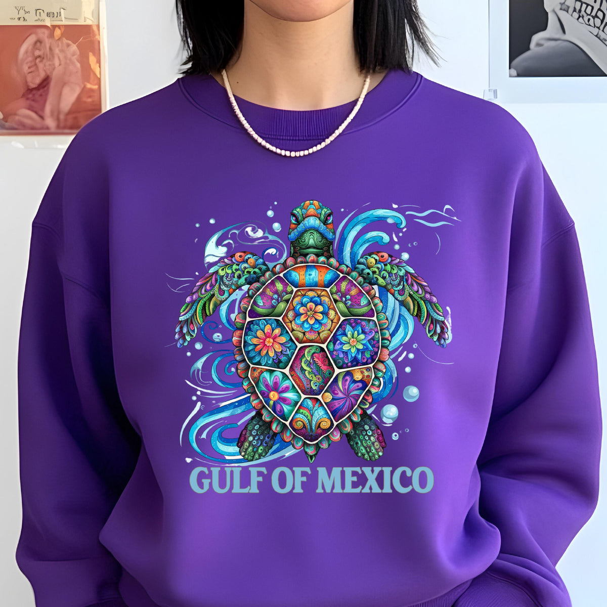 May include: A purple sweatshirt featuring a colorful sea turtle design. The turtle's shell is decorated with floral patterns in various colors. The words "GULF OF MEXICO" are printed in light blue below the turtle. The sweatshirt has a crew neck.