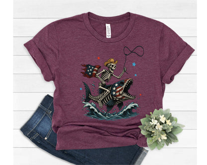 May include: Maroon t-shirt featuring a skeleton riding a fish with an American flag design. The skeleton wears a cowboy hat and holds a lasso. The fish has an American flag pattern, with blue waves and red and blue stars.