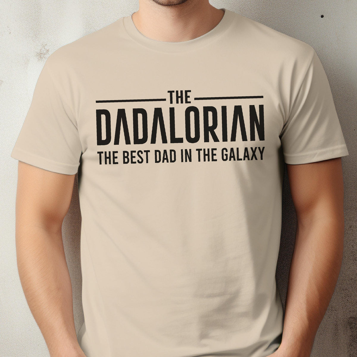 May include: Beige t-shirt with the text "THE DADALORIAN THE BEST DAD IN THE GALAXY" in black. The shirt is a crew neck style and appears to be made of a soft material. The text is centered on the front of the shirt.
