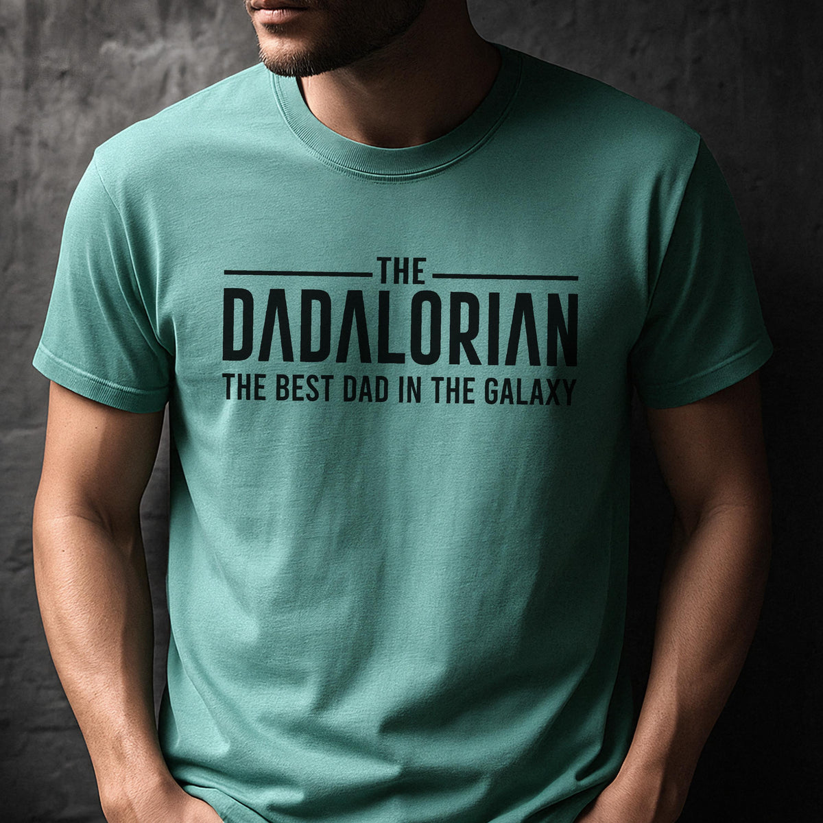 May include: A teal t-shirt with the text "THE DADALORIAN THE BEST DAD IN THE GALAXY" in black. The shirt has short sleeves and a crew neck. The text is centered on the chest.