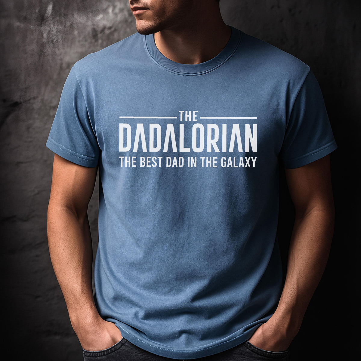 May include: A light blue t-shirt with the text "THE DADALORIAN THE BEST DAD IN THE GALAXY" in white. The shirt has short sleeves and a crew neck. The person wearing the shirt has their hands in their pockets.