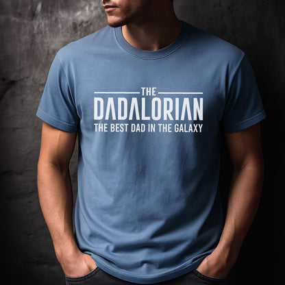 May include: A light blue t-shirt with the text "THE DADALORIAN THE BEST DAD IN THE GALAXY" in white. The shirt has short sleeves and a crew neck. The person wearing the shirt has their hands in their pockets.