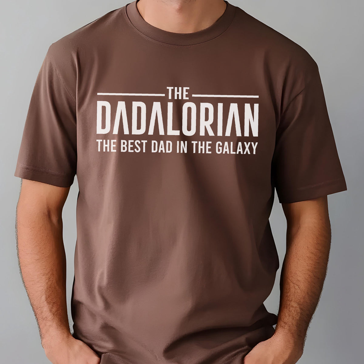 May include: Brown t-shirt with the text "THE DADALORIAN THE BEST DAD IN THE GALAXY" in white. The shirt has a crew neck and short sleeves. The text is centered on the front of the shirt.