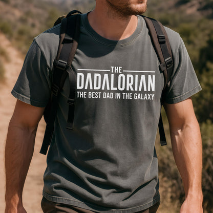 May include: Dark gray t-shirt with the text "THE DADALORIAN THE BEST DAD IN THE GALAXY" in white. The shirt is short-sleeved and has a crew neck. The person wearing the shirt is also wearing a black backpack.