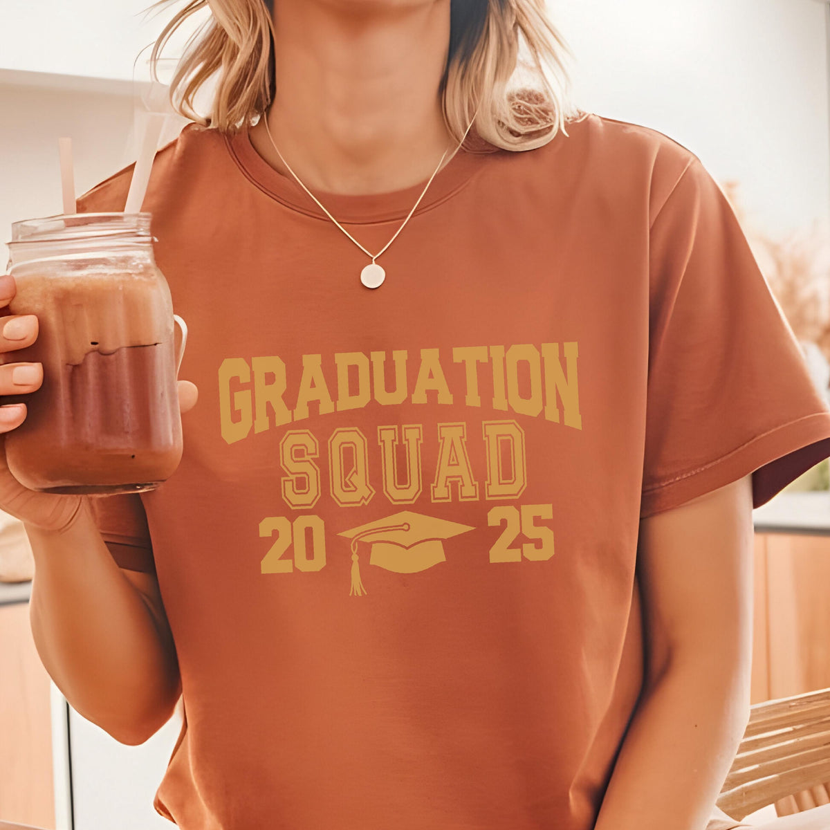 May include: A rust-colored t-shirt with the words "GRADUATION SQUAD 2025" in a gold, block-letter font. A graduation cap graphic is below the text. The shirt is short-sleeved.