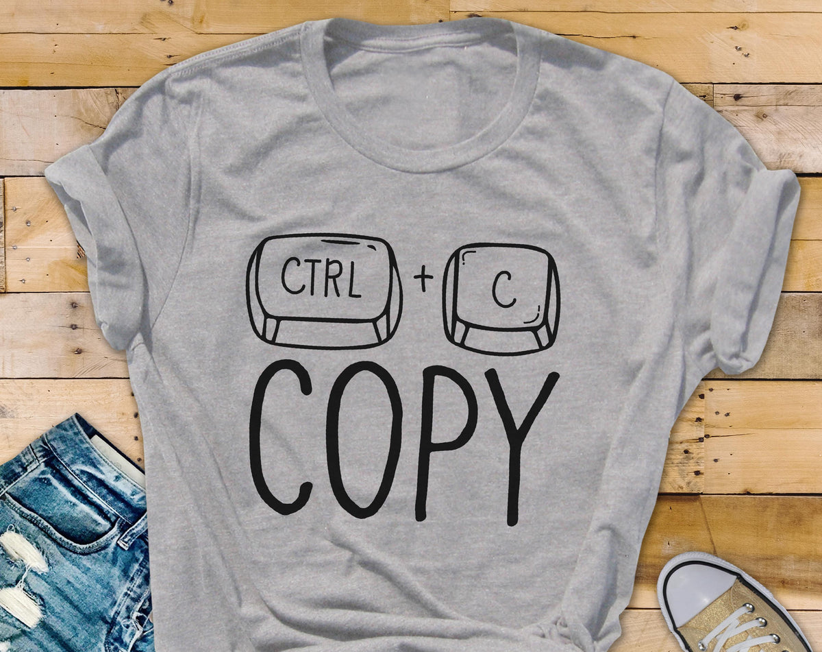 May include: A light gray t-shirt with a black graphic of the CTRL and C keys, with the word "COPY" below. The shirt is made of a soft material and is suitable for casual wear. A pair of blue jeans and a sneaker are visible.