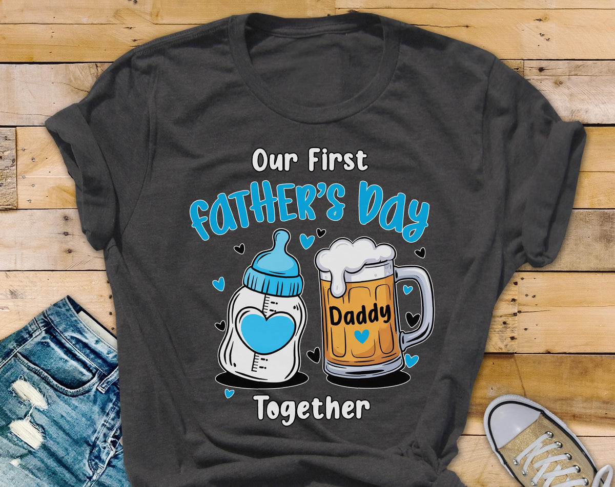 May include: Dark gray t-shirt with the text "Our First Father's Day Together." The design includes a baby bottle with a blue heart and a beer mug with the word "Daddy." Blue hearts are scattered around the design.