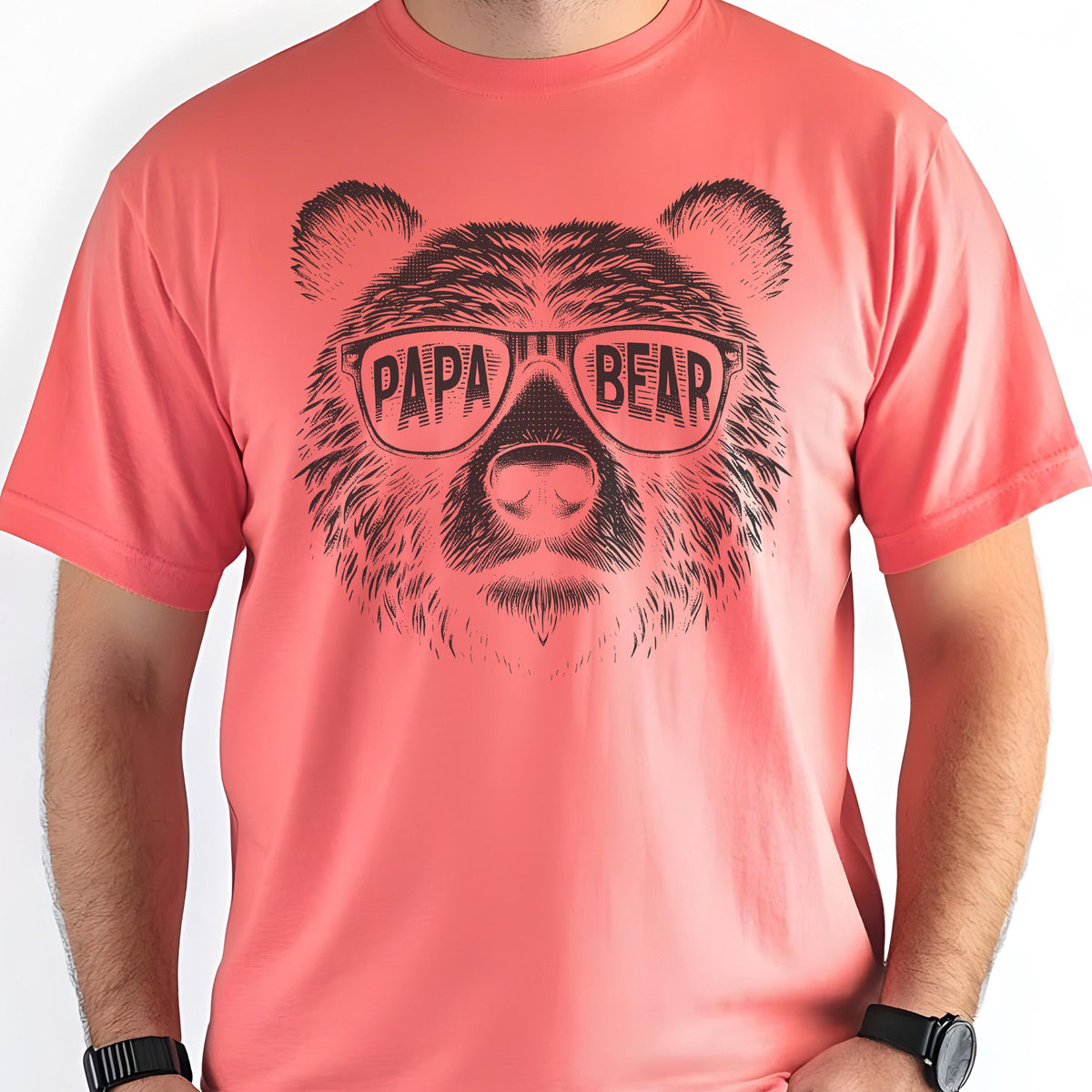 May include: Coral-colored t-shirt featuring a black graphic of a bear's face wearing sunglasses. The sunglasses have the words "PAPA BEAR" printed on them. The shirt is short-sleeved and made of a soft material.