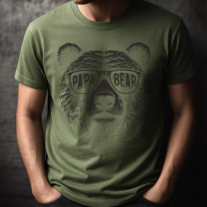 May include: Olive green t-shirt featuring a detailed illustration of a bear's face wearing glasses. The glasses have the words "PAPA BEAR" printed on them in a bold, sans-serif font. The shirt is a solid color and has short sleeves.