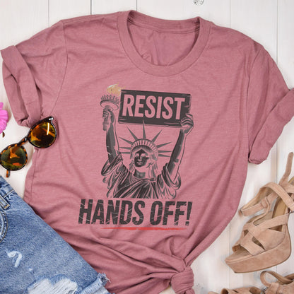 May include: A pink t-shirt with a graphic of the Statue of Liberty holding a sign that says "Resist" and the text "Hands Off!" printed below.