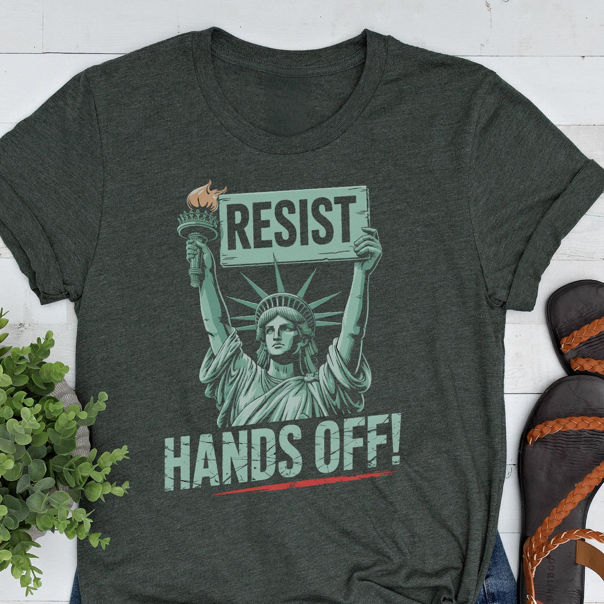 May include: A dark green t-shirt with a graphic of the Statue of Liberty holding a sign that says "Resist" and the text "Hands Off!" printed below the graphic.