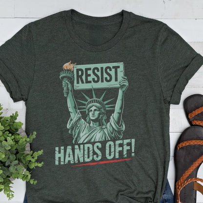 May include: A dark green t-shirt with a graphic of the Statue of Liberty holding a sign that says "Resist" and the text "Hands Off!" printed below the graphic.
