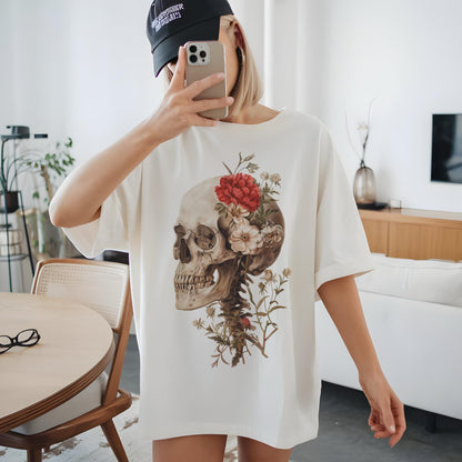 May include: Cream-colored t-shirt featuring a detailed skull graphic adorned with red and white flowers. The design includes the skull's jaw and spine, with floral accents. The shirt is a casual, oversized fit.