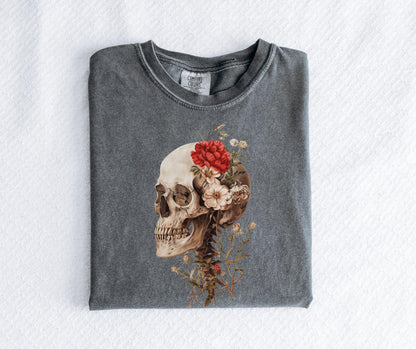 May include: A gray Comfort Colors t-shirt featuring a detailed illustration of a human skull adorned with red and cream-colored flowers. The design includes the spinal column and additional floral elements.