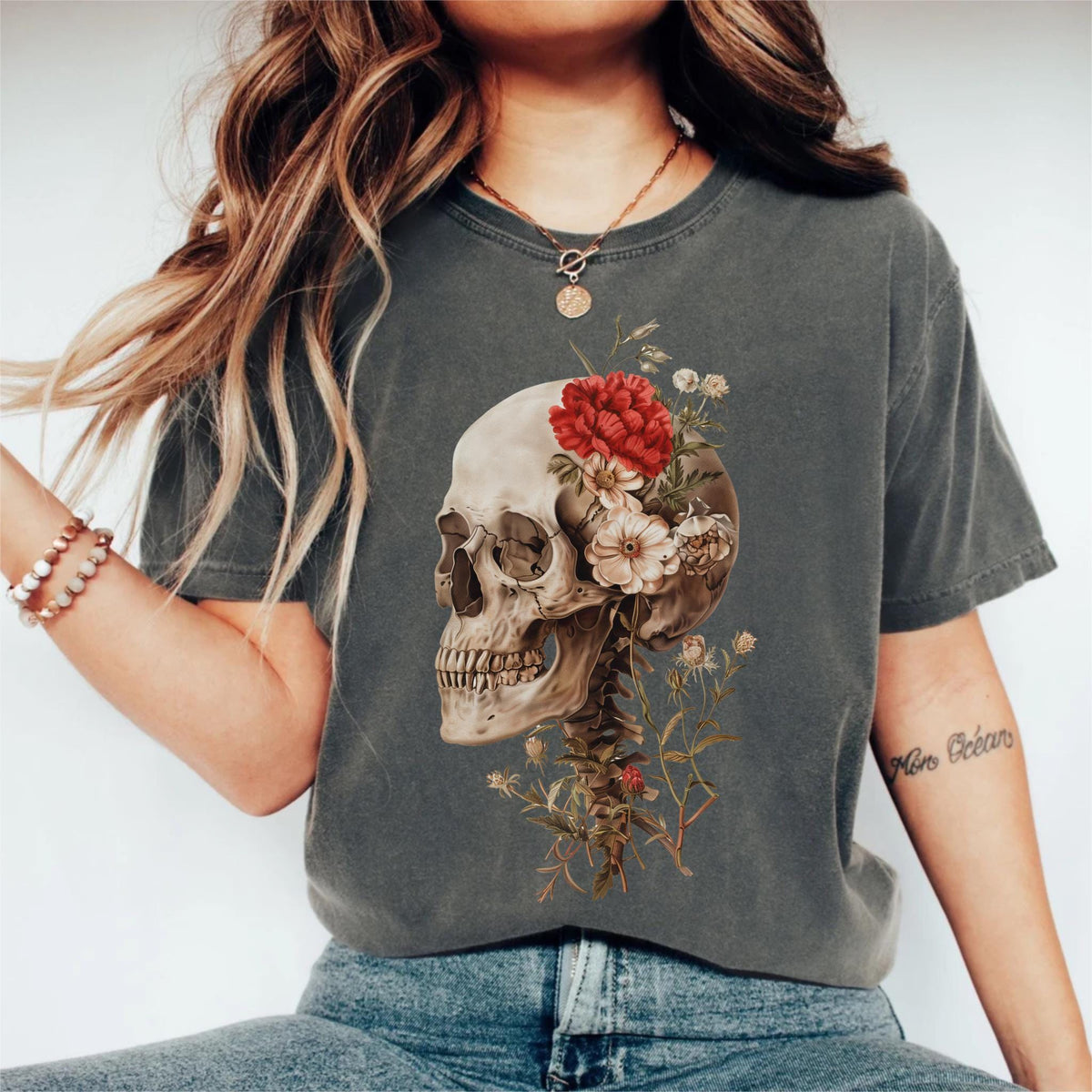May include: Dark gray t-shirt featuring a detailed skull graphic adorned with red and white flowers. The design includes a spine and floral elements, creating a unique and artistic aesthetic. The shirt is a comfortable, casual wear.