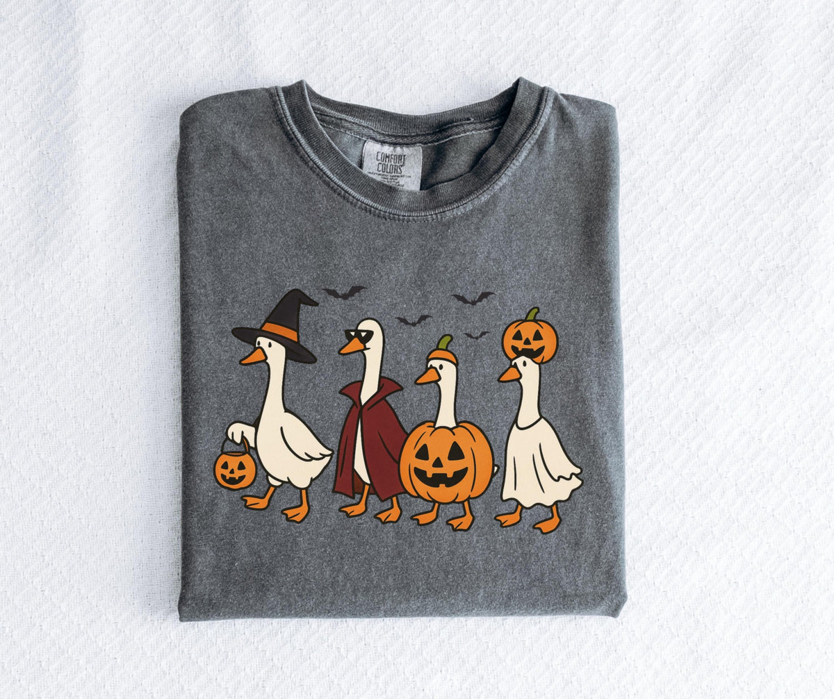 May include: Dark grey Comfort Colors t-shirt featuring a Halloween-themed graphic. The design shows four cartoon geese dressed in costumes: a witch, a vampire, a jack-o'-lantern, and a ghost. Bats fly above them.