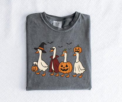 May include: Dark grey Comfort Colors t-shirt featuring a Halloween-themed graphic. The design shows four cartoon geese dressed in costumes: a witch, a vampire, a jack-o'-lantern, and a ghost. Bats fly above them.