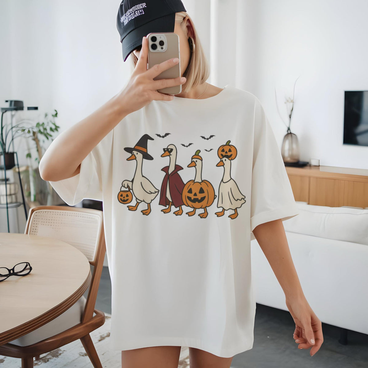 May include: Cream-colored t-shirt featuring a Halloween-themed graphic. The design showcases four cartoon geese dressed in Halloween costumes: a witch, a vampire, a jack-o'-lantern, and a ghost. Bats fly above the geese. The shirt is a casual, oversized fit.