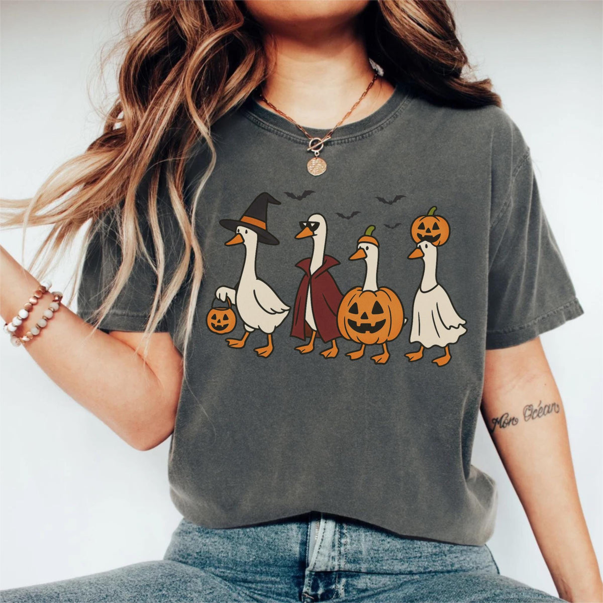 May include: Dark gray t-shirt featuring a Halloween-themed graphic. The design shows four cartoon geese dressed in costumes: a witch, a vampire, a pumpkin, and a ghost. The geese are walking in a row, with bats flying above them.