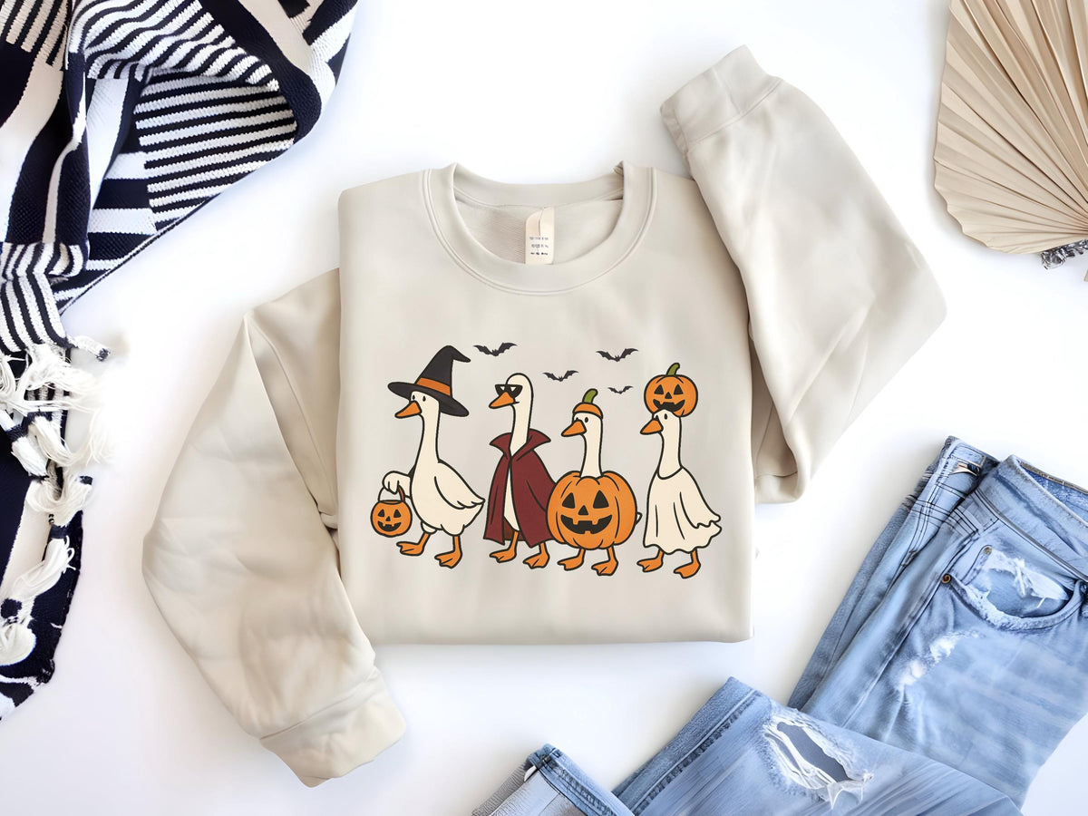 May include: Beige sweatshirt featuring a Halloween-themed graphic of four geese in costumes: a witch, a vampire, a jack-o'-lantern, and a ghost. The geese are walking, with bats flying above them. The sweatshirt is paired with blue jeans.