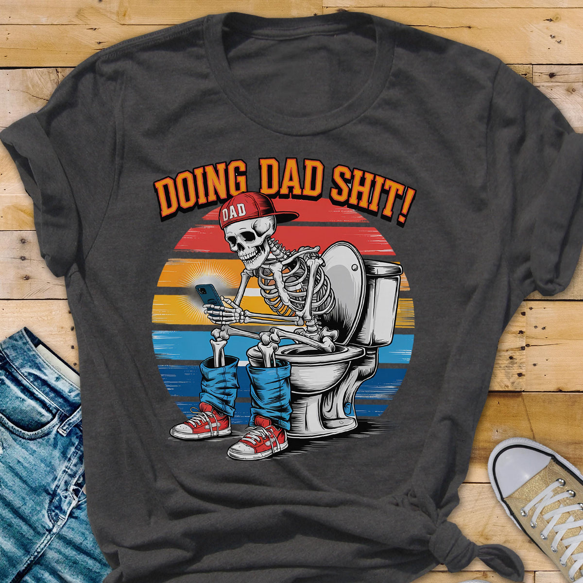 May include: Dark gray t-shirt with a humorous graphic. The design features a skeleton wearing a red cap, blue jeans, and red sneakers, sitting on a toilet while using a smartphone. The text "DOING DAD SHIT!" is above the illustration.