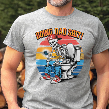 May include: Gray t-shirt with a graphic of a skeleton wearing a red cap and blue jeans, sitting on a toilet while using a smartphone. The text "DOING DAD SHIT!" is above the skeleton, with "DAD" on the cap.