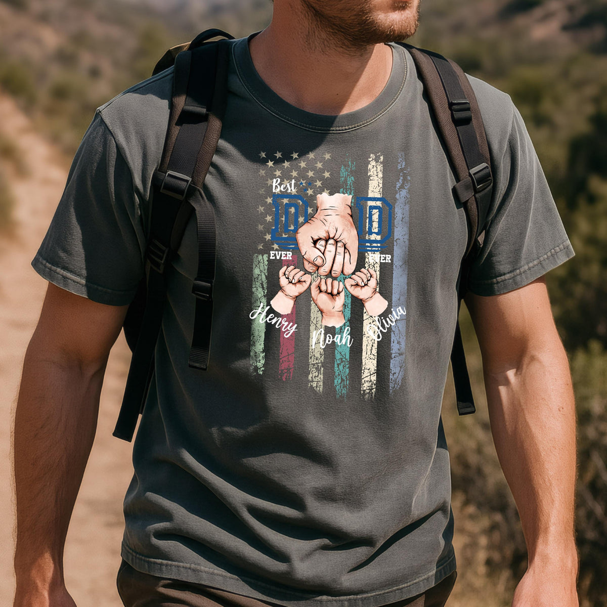 May include: Dark gray t-shirt featuring a distressed American flag design with the words "Best Dad Ever." The graphic includes a father's fist and three smaller fists with the names Henry, Noah, and Olivia. The shirt is worn by a person with a backpack.