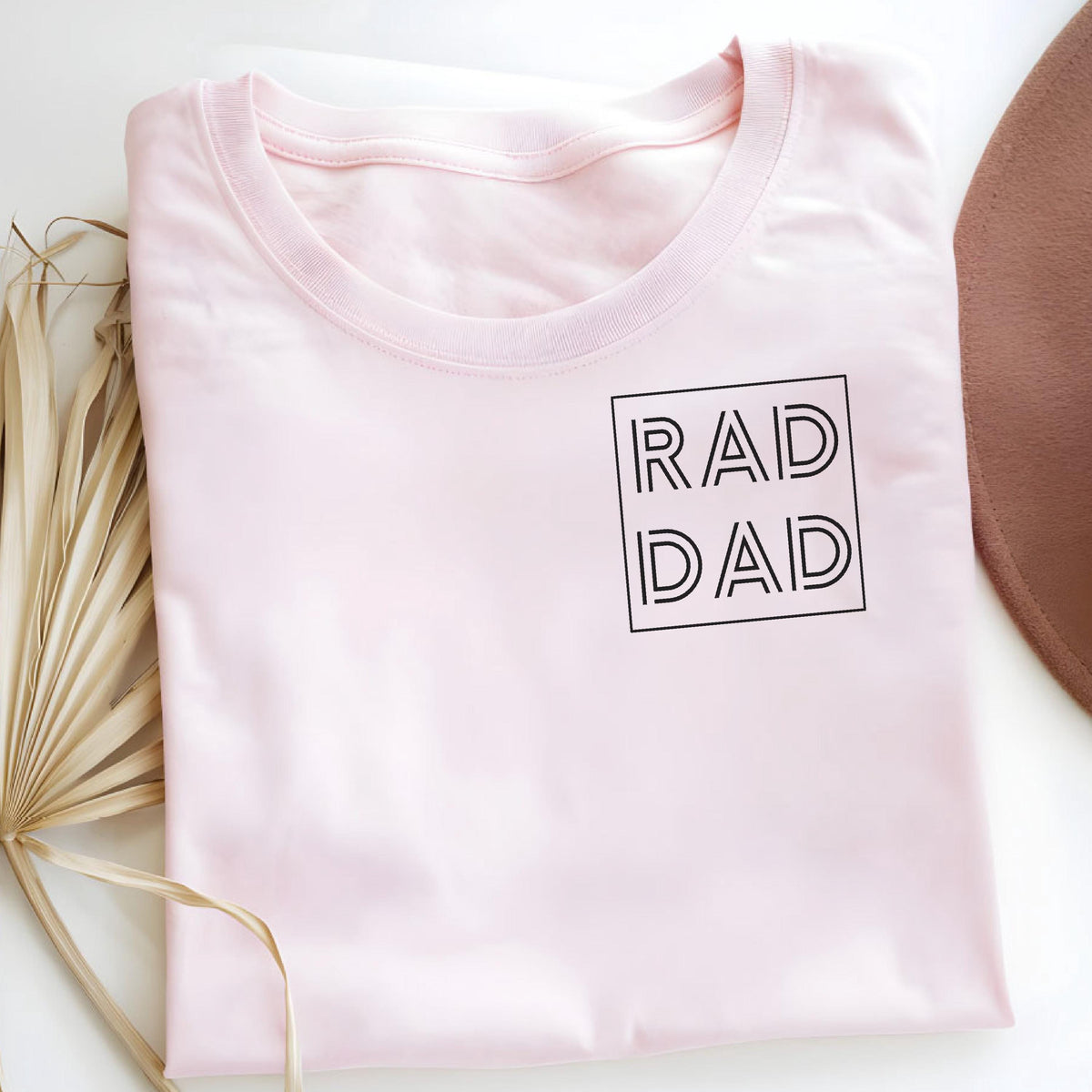 May include: A light pink t-shirt with the words "RAD DAD" in a black, outlined font within a square. The shirt is folded and lies on a white surface, with a brown hat and dried palm leaves in the background.