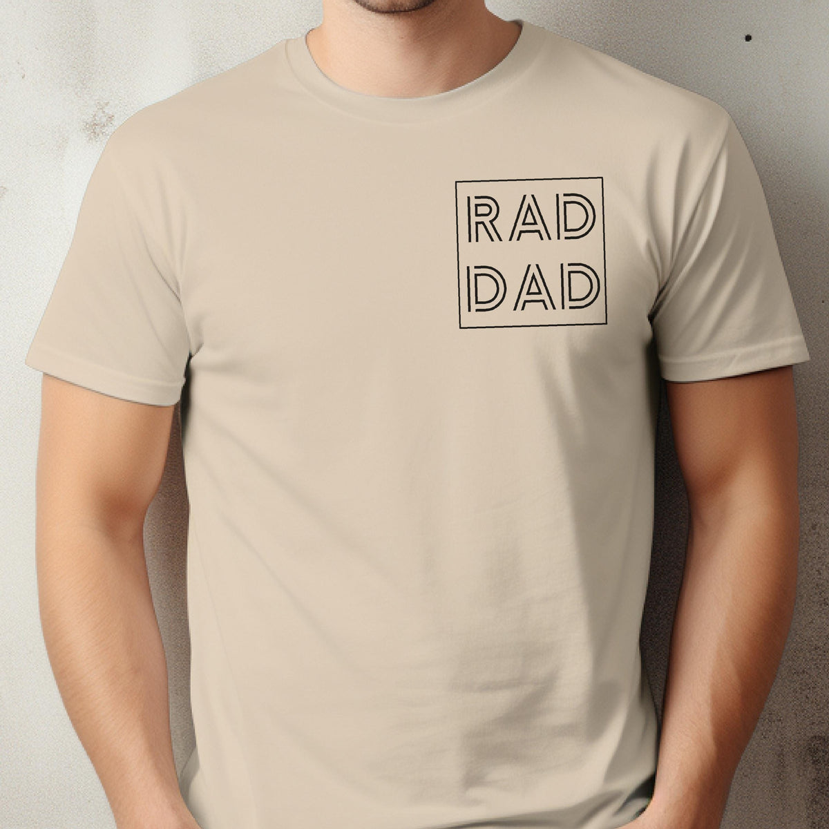 May include: Beige t-shirt with the text "RAD DAD" in a black, square design on the upper left chest. The shirt has short sleeves and a crew neck. The text is in a bold, sans-serif font.