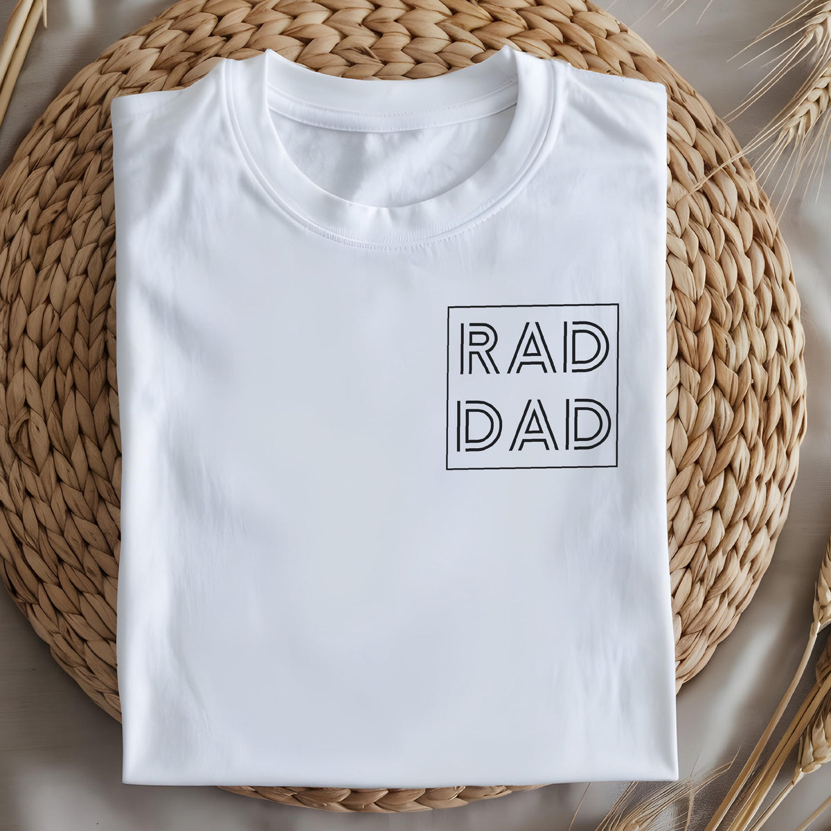 May include: White t-shirt with the text "RAD DAD" in a black, minimalist font, enclosed in a square. The shirt is neatly folded on a woven, light brown surface, with wheat stalks in the background. A casual, everyday apparel item.
