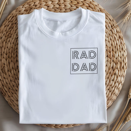 May include: White t-shirt with the text "RAD DAD" in a black, minimalist font, enclosed in a square. The shirt is neatly folded on a woven, light brown surface, with wheat stalks in the background. A casual, everyday apparel item.