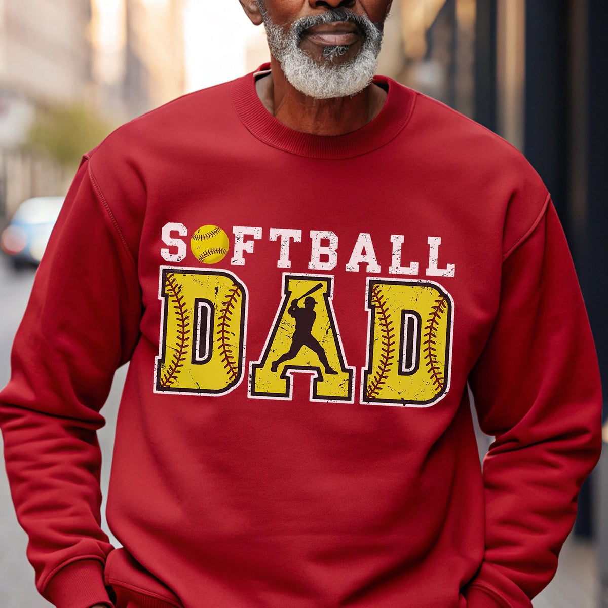 May include: A red sweatshirt with the words "SOFTBALL DAD" in white and yellow. The word "SOFTBALL" is above the word "DAD". The letter "D" in "DAD" has a silhouette of a batter swinging a bat.