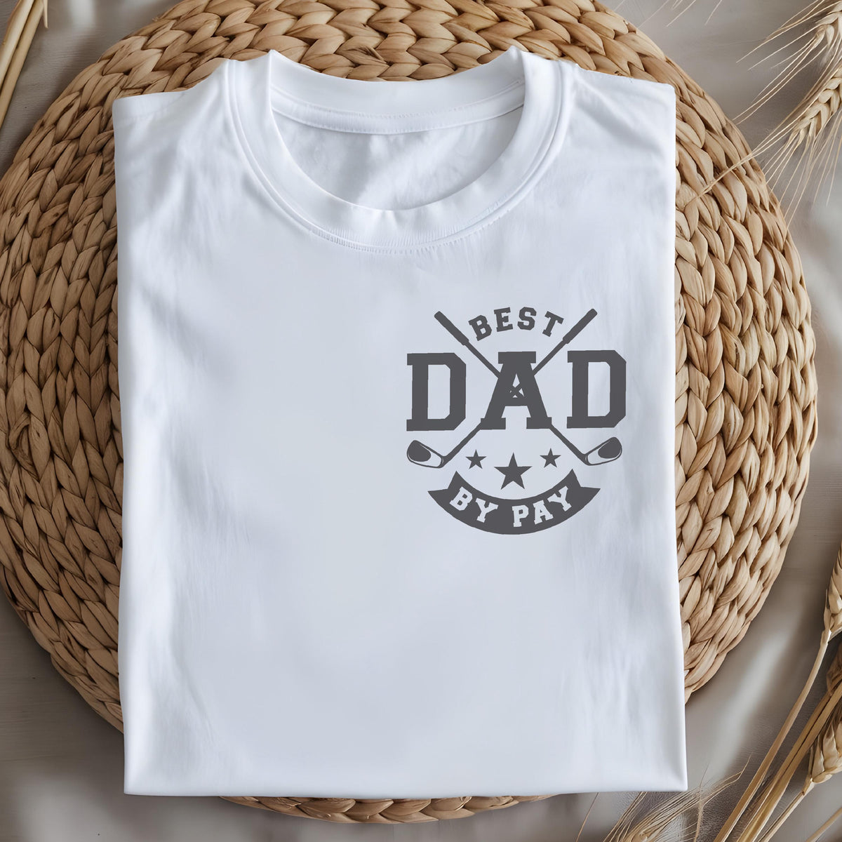 May include: White t-shirt with a crew neck and a gray graphic. The graphic reads "BEST DAD BY PAY" with crossed golf clubs and stars. The shirt is folded neatly on a woven surface.