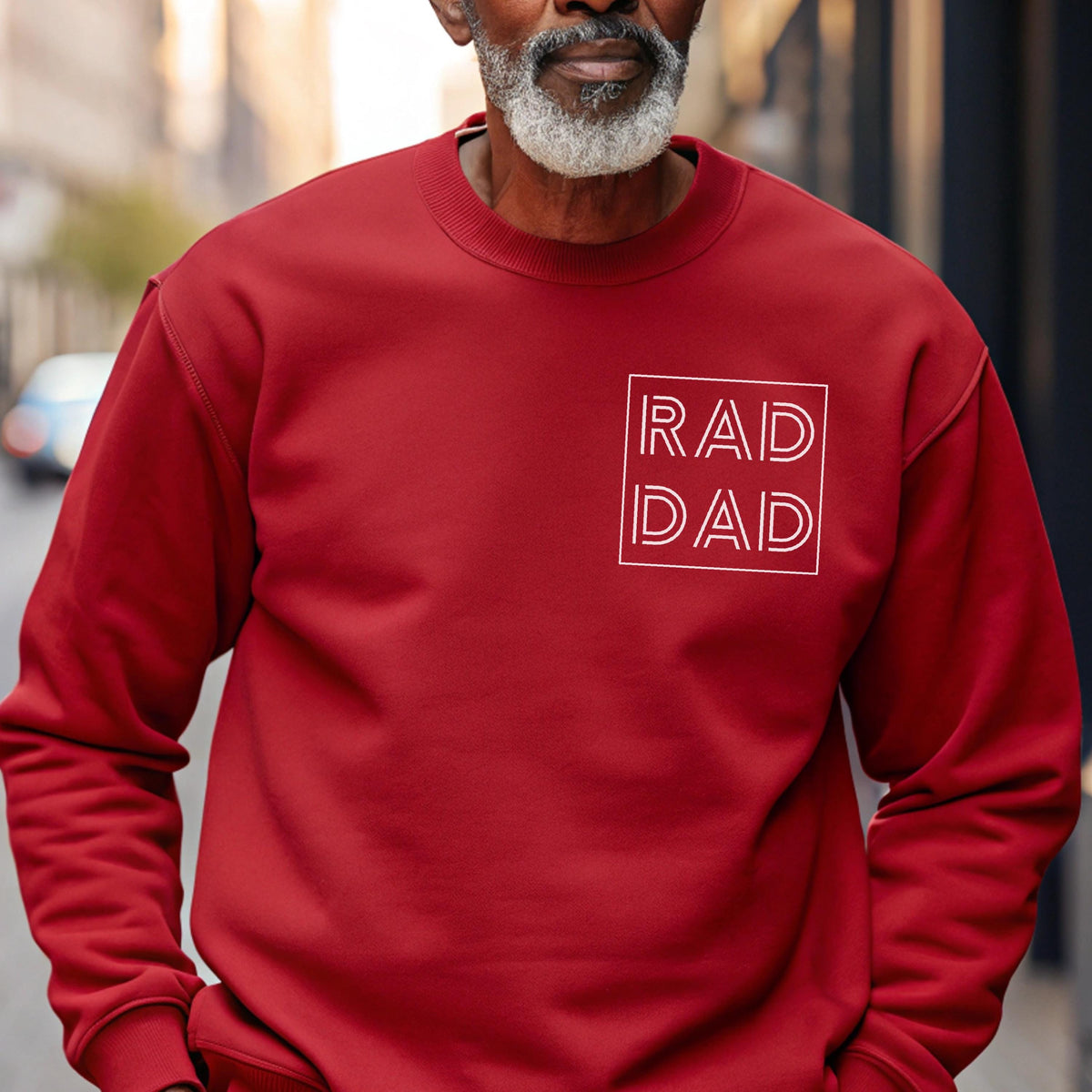 May include: A red sweatshirt with the words "RAD DAD" in a white square. The sweatshirt is a solid color and has a crew neck. The text is in a simple, sans-serif font.