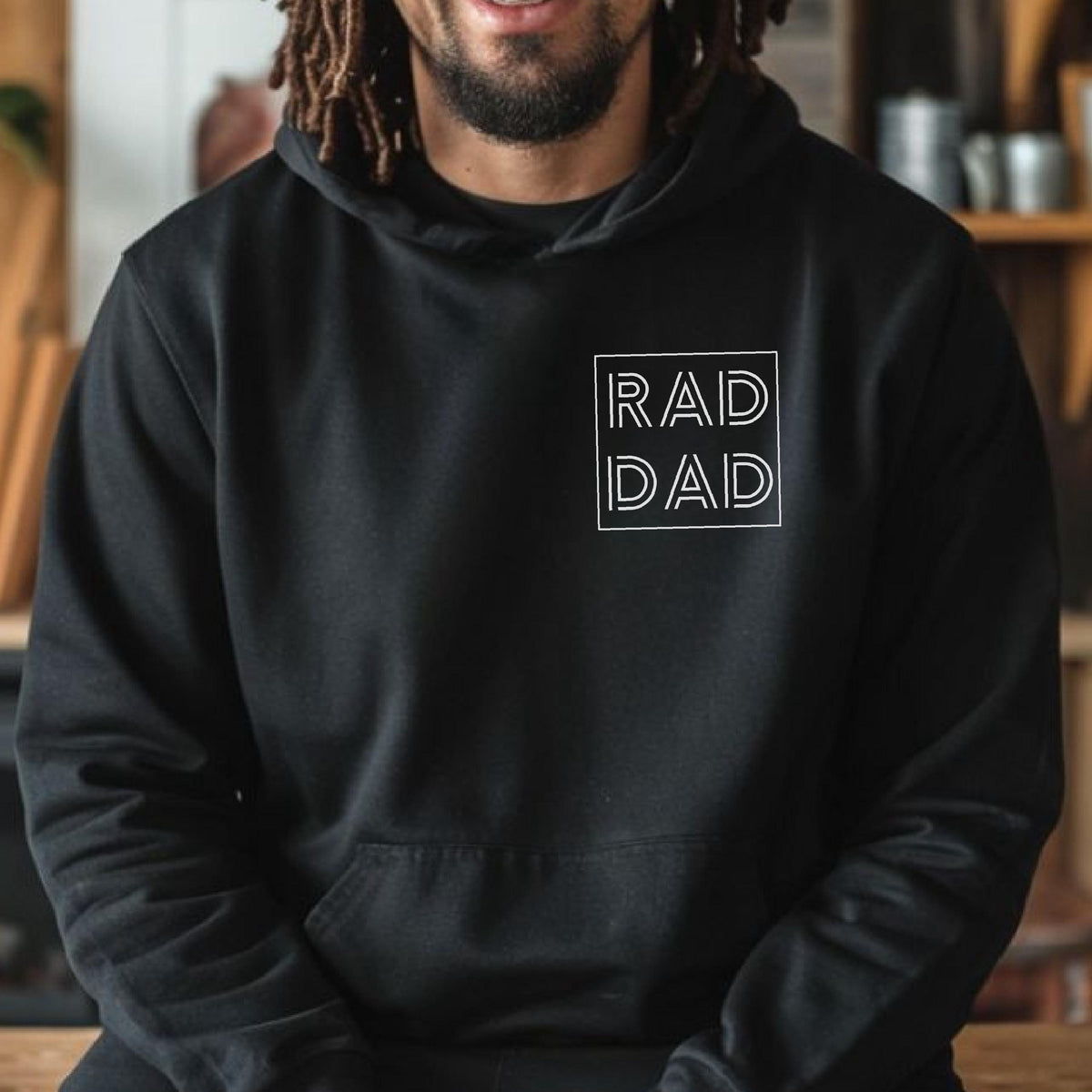 May include: Black hoodie with a front pocket and a white square design. The design features the words "RAD DAD" in a minimalist font. The hoodie has a hood and long sleeves.