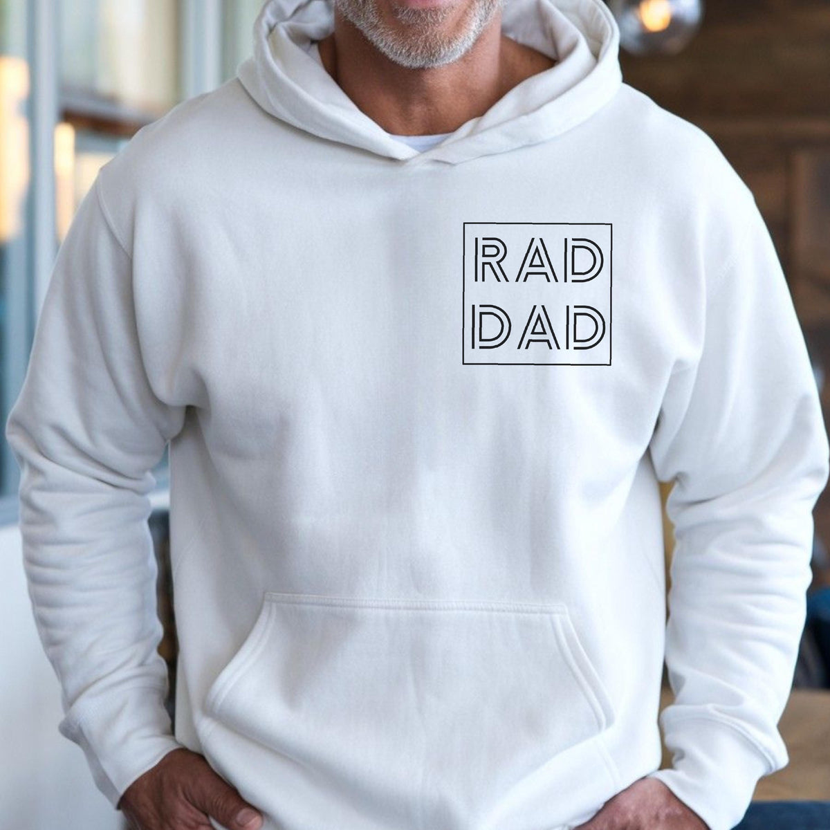 May include: A white hoodie with a front pocket and a drawstring hood. The hoodie features the text "RAD DAD" in a black, minimalist font within a square frame on the chest. The garment is made of a soft material.