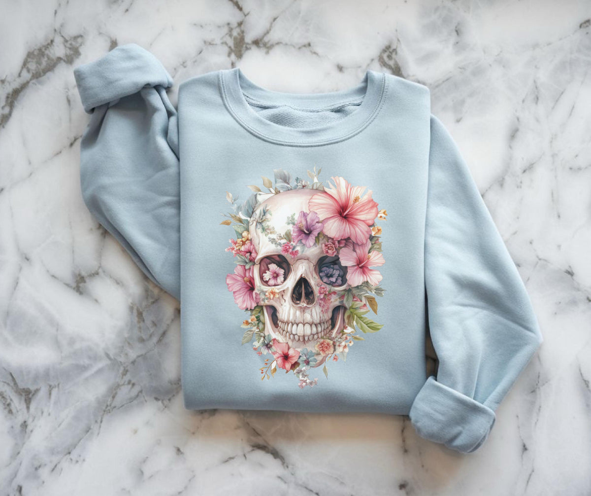 May include: A light blue sweatshirt featuring a watercolor skull design adorned with pink, purple, and white flowers. The skull's eye sockets are filled with blue and purple hues, and the design is centered on the front of the garment.