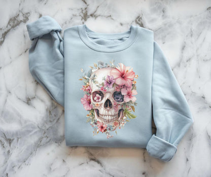 May include: A light blue sweatshirt featuring a watercolor skull design adorned with pink, purple, and white flowers. The skull's eye sockets are filled with blue and purple hues, and the design is centered on the front of the garment.