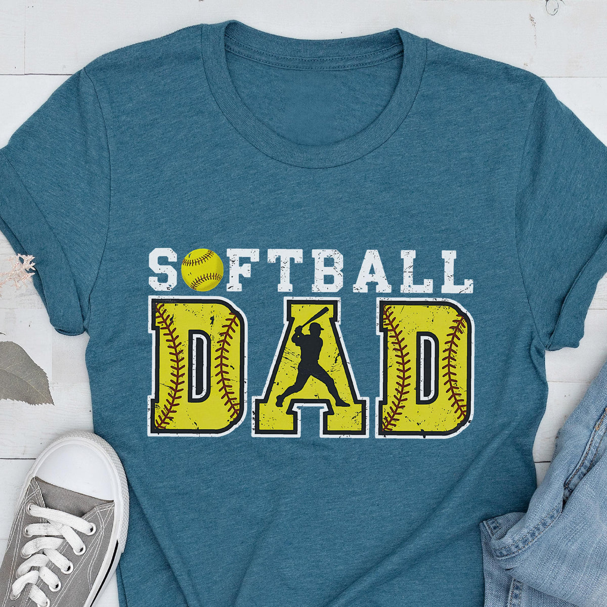 May include: Teal blue t-shirt with the text "Softball Dad" in white with a yellow softball and a silhouette of a softball player in the middle of the word "Dad".