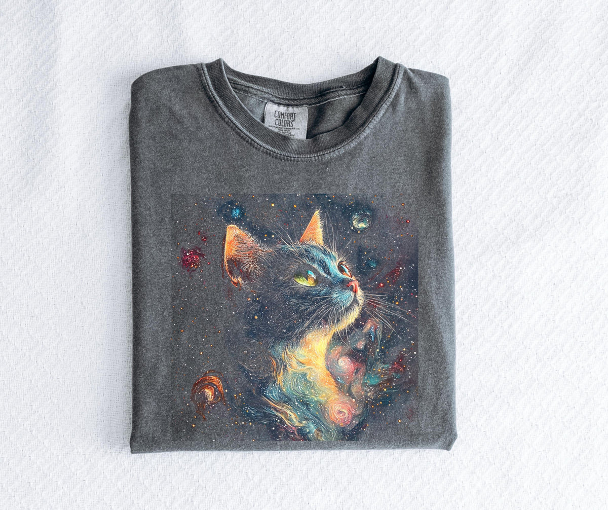 May include: A gray Comfort Colors t-shirt featuring a colorful illustration of a cat with a galaxy design. The cat has yellow eyes and orange ears, set against a dark background with stars and planets. The shirt is folded neatly on a white surface.