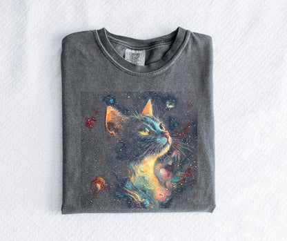 May include: A gray Comfort Colors t-shirt featuring a colorful illustration of a cat with a galaxy design. The cat has yellow eyes and orange ears, set against a dark background with stars and planets. The shirt is folded neatly on a white surface.