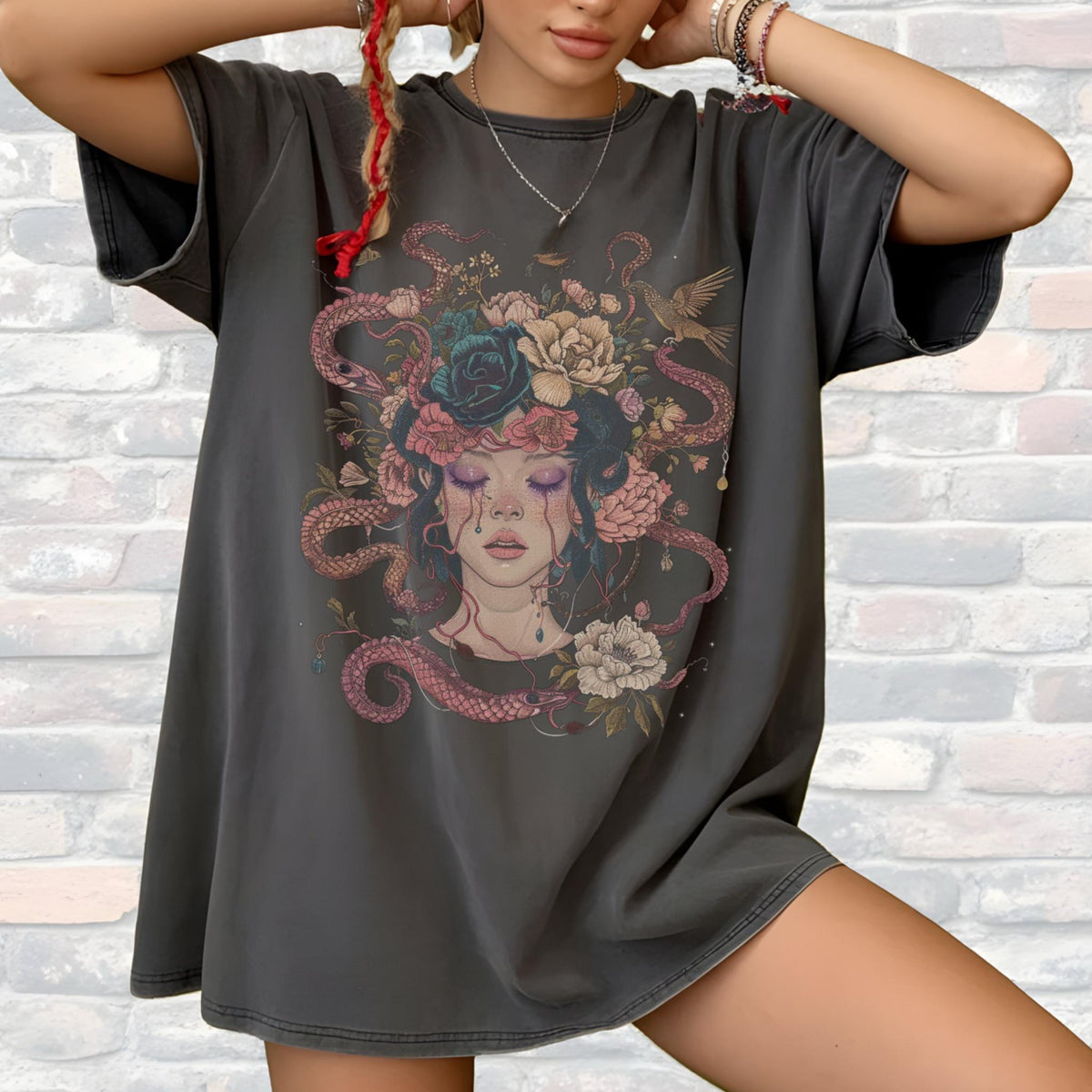 May include: Dark gray oversized t-shirt featuring a detailed illustration of a woman with a floral and snake headdress. The artwork includes roses, snakes, and a bird, with a color palette of pink, blue, and gray. The shirt has short sleeves.