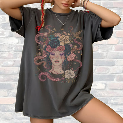 May include: Dark gray oversized t-shirt featuring a detailed illustration of a woman with a floral and snake headdress. The artwork includes roses, snakes, and a bird, with a color palette of pink, blue, and gray. The shirt has short sleeves.