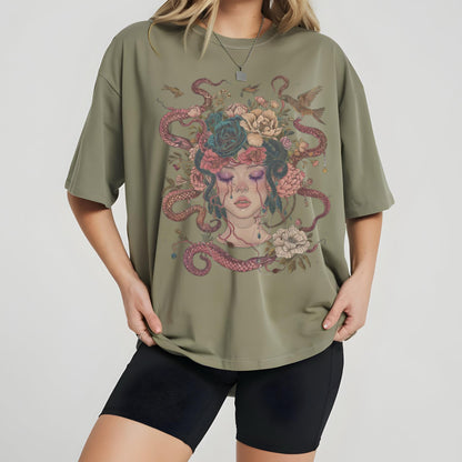 May include: Olive green t-shirt featuring a detailed illustration of a woman with snakes for hair, adorned with flowers and birds. The artwork has a vintage, artistic style. The shirt is paired with black shorts.