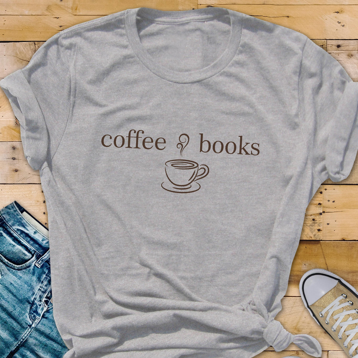 May include: A heather gray t-shirt with the words "coffee & books" in brown script above a coffee cup illustration. The shirt is knotted at the waist and paired with blue jeans and a canvas shoe.