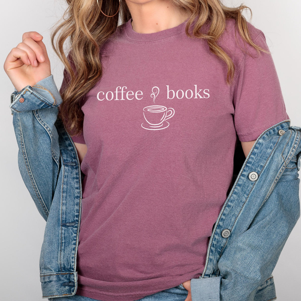 May include: A mauve short-sleeved t-shirt featuring the white text coffee & books with a line art graphic of a steaming coffee cup. The shirt is worn under a blue denim jacket with metal buttons, showcasing a casual and layered appearance.