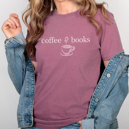 May include: A mauve short-sleeved t-shirt featuring the white text coffee & books with a line art graphic of a steaming coffee cup. The shirt is worn under a blue denim jacket with metal buttons, showcasing a casual and layered appearance.